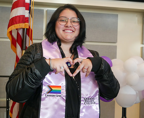 A lavender graduate wears her sash and makes a heart with her fingers