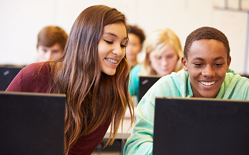 Two students in a classroom work on laptops and collaborate on an assignment