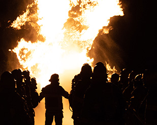 Firefighter trainees stand silhouetted against a plume of fire during training