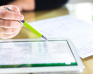 Closeup of a hand holding a pen and a tablet screen showing a spreadsheet