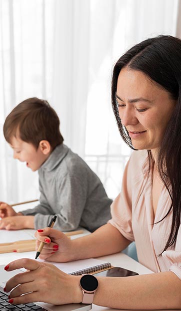 An adult woman works on school work while her son plays in the background