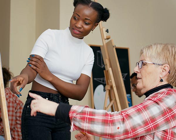 A young art instructor gives tips to an older adult student drawing at an easel