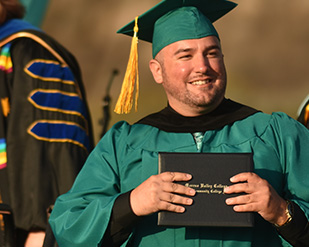 A student in teal graduation regalia holds up their degree holder at Commencement