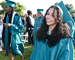 A grinning MVC graduate walks onto the Commencement field in teal regalia
