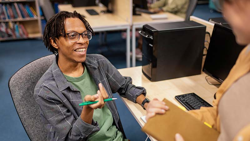 A student talks to another while sitting at a computer in the library