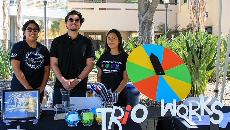 Three students stand behind a table loaded with TRIO activities and information