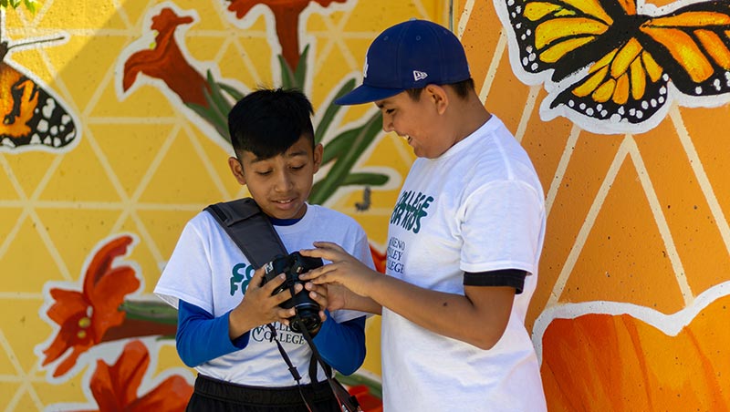 Two program participants use a digital camera in front of MVC's campus mural