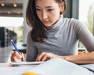 A student writes in a notebook while looking at her textbook