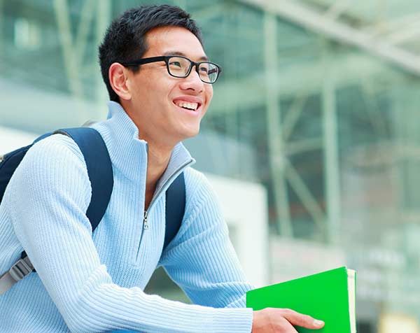 A grinning young man sits in front of a university building with a backpack and notebook