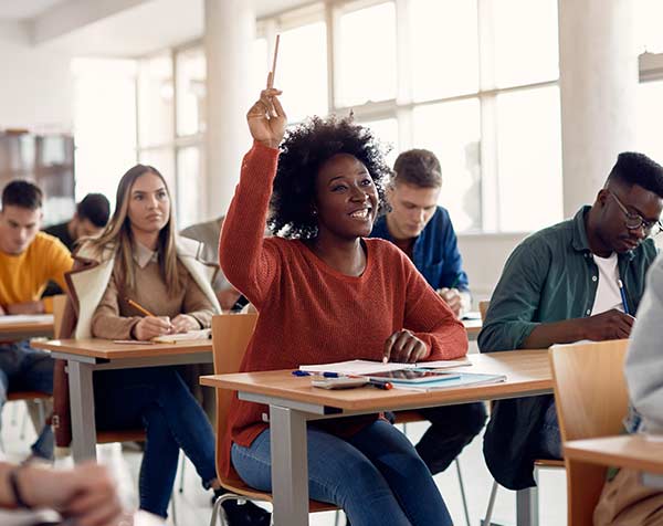 A student, in a classroom surrounded by other young adults, raises her hand to answer a question