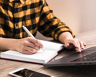 A student holds a pencil while working in a notebook next to a phone and laptop