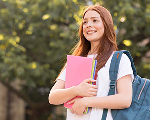 A student grins while holding folders with a backpack hanging off one shoulder