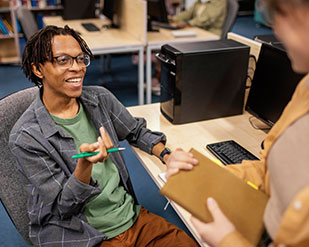 A student lounges in a computer chair while talking to another student standing in front of them